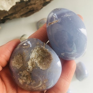 May include: Two smooth, oval-shaped blue chalcedony stones. One stone has a rough, textured area with brown and beige tones. The other stone is a solid, pale blue color. Additional stones are visible in the background.
