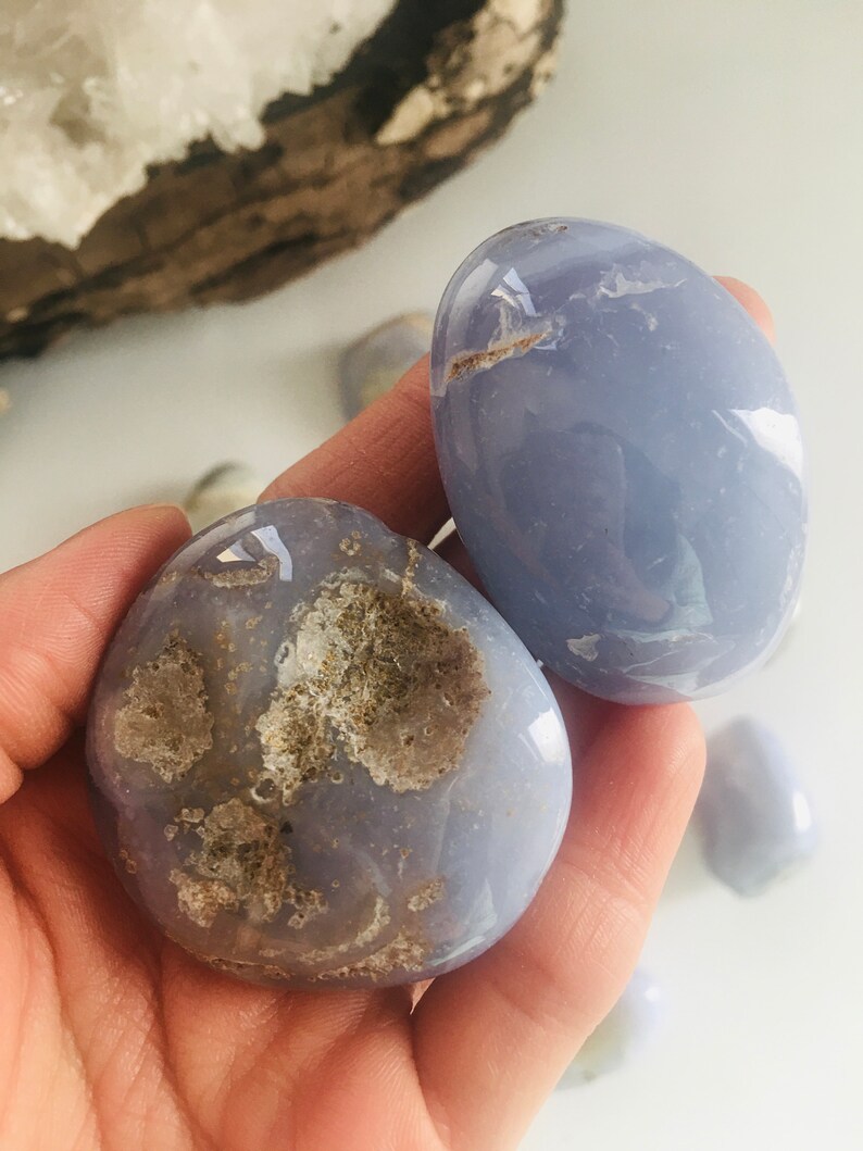 May include: Two smooth, oval-shaped blue chalcedony stones. One stone has a rough, textured area with brown and beige tones. The other stone is a solid, pale blue color. Additional stones are visible in the background.