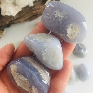 May include: Several polished blue lace agate stones with white and tan inclusions. The stones are smooth and rounded, with a soft, translucent appearance. The stones are held in a hand, with other stones visible in the background. The stones are a light blue color.