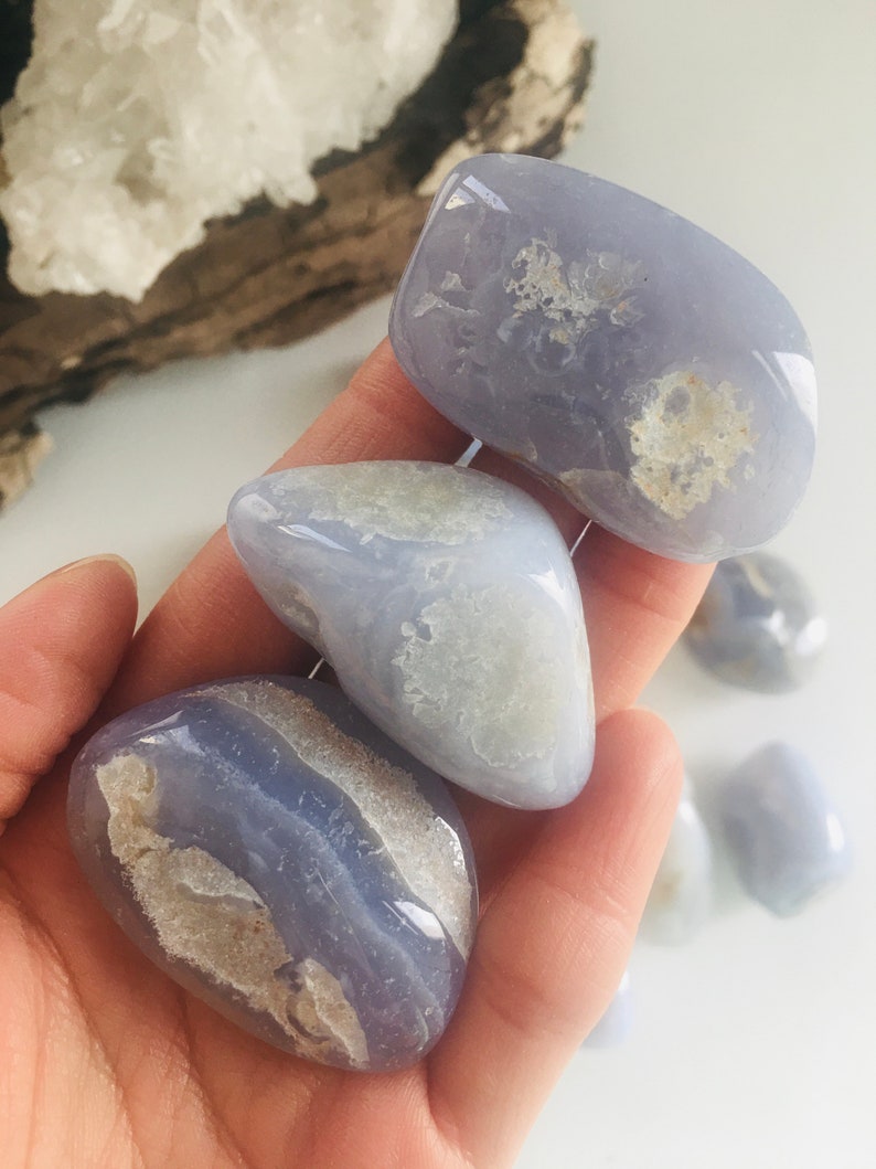 May include: Several polished blue lace agate stones with white and tan inclusions. The stones are smooth and rounded, with a soft, translucent appearance. The stones are held in a hand, with other stones visible in the background. The stones are a light blue color.
