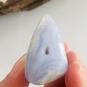 May include: A polished, light blue gemstone with a smooth, rounded shape. The stone has a small, dark cavity and a swirling pattern of lighter blue and white. The gemstone is held in a person's hand.