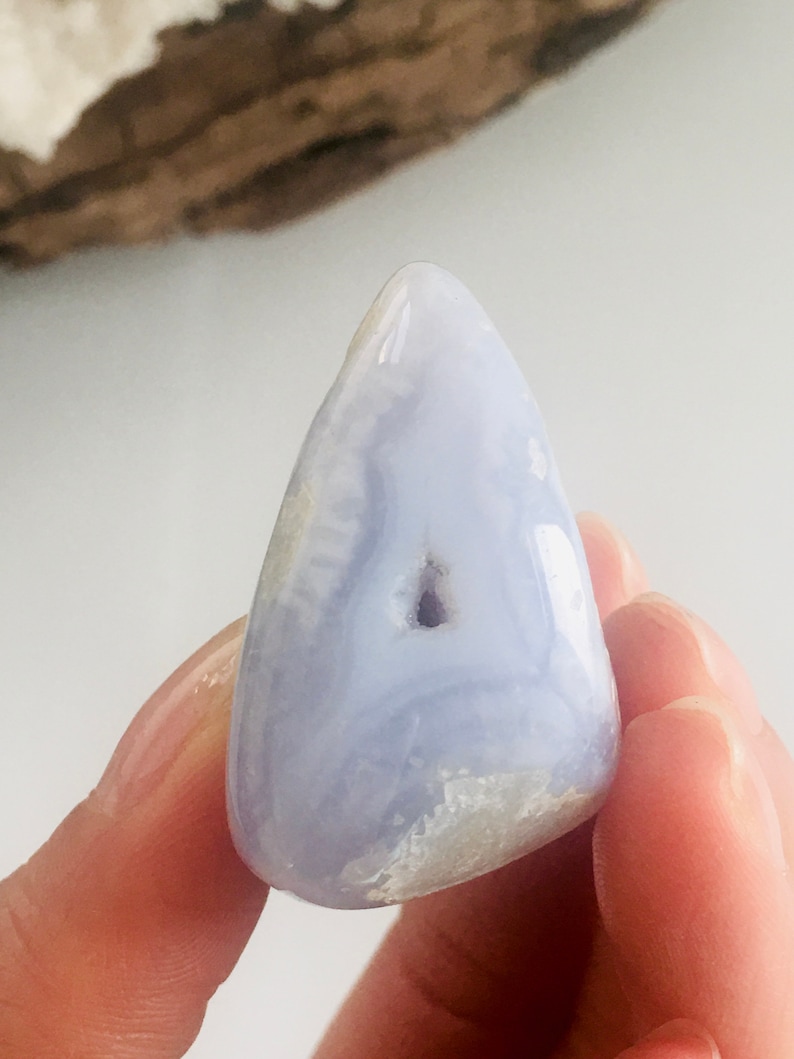 May include: A polished, light blue gemstone with a smooth, rounded shape. The stone has a small, dark cavity and a swirling pattern of lighter blue and white. The gemstone is held in a person's hand.