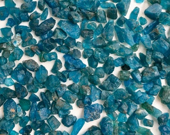 May include: A collection of blue apatite crystal chips. The chips are various sizes and shapes, with some having a more pointed shape. The chips are scattered on a white background.