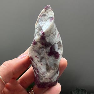 May include: A polished, natural gemstone with a unique shape. The stone features a mix of white, gray, and deep red hues. The surface is smooth and reflective, with visible patterns and variations in color. A decorative item.