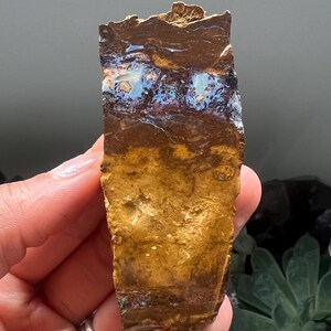 Könnte beinhalten: Ein rohes Boulder-Opal-Exemplar mit einer dunkelbraunen Eisensteinmatrix, einem horizontalen Band aus irisierenden blauen und grünen Opal-Blitzen im oberen Bereich und einer texturierten, erdig gelb-braunen Basis mit natürlichen Mineraleinschlüssen.