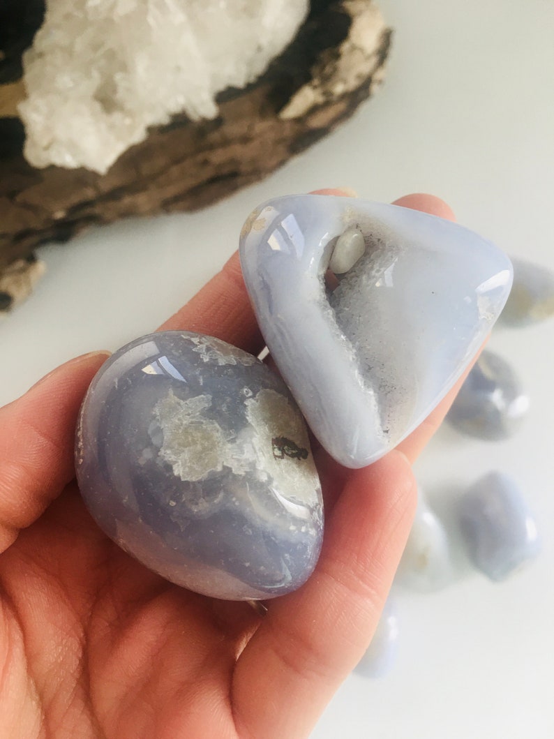 May include: Two smooth, blue lace agate stones are held in a hand. The stones have a translucent appearance with white and gray inclusions. The background includes additional stones and a piece of driftwood.