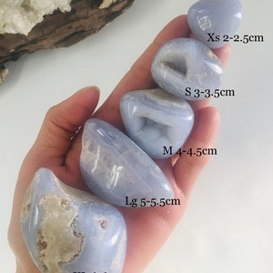 May include: A hand holds a collection of polished blue lace agate stones in various sizes, labeled from XS to XL, with measurements in centimeters. The stones have a pale blue hue with white and tan inclusions, and some have small cavities.