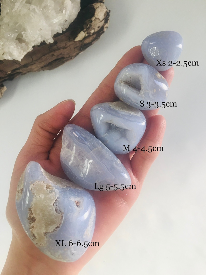 May include: A hand holds a collection of polished blue lace agate stones in various sizes, labeled from XS to XL, with measurements in centimeters. The stones have a pale blue hue with white and tan inclusions, and some have small cavities.