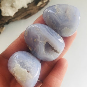 May include: Three smooth, light blue chalcedony stones with white and beige inclusions are held in a hand. The stones have a polished, rounded shape. The background includes a piece of wood and a white surface.