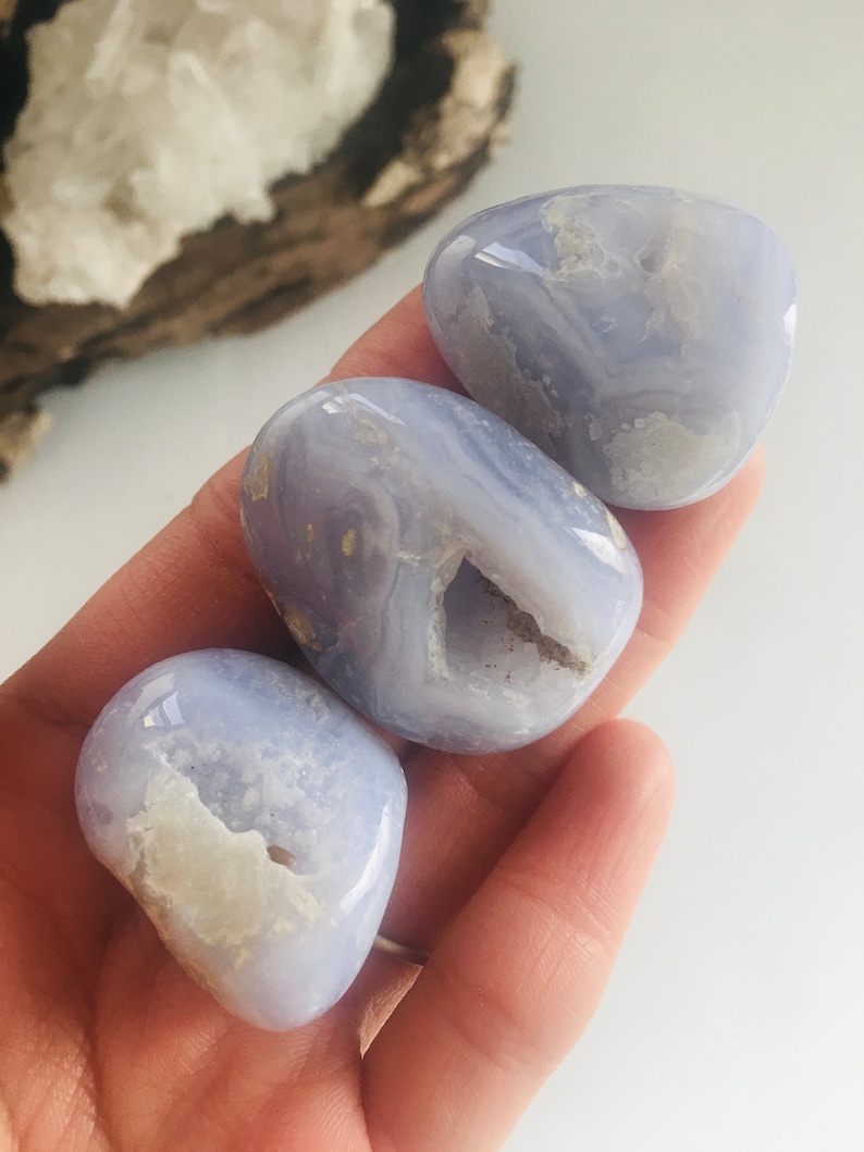 May include: Three smooth, light blue chalcedony stones with white and beige inclusions are held in a hand. The stones have a polished, rounded shape. The background includes a piece of wood and a white surface.