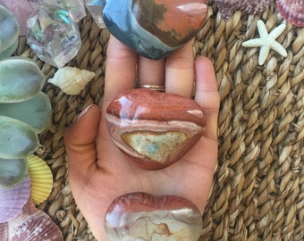 May include: Three heart-shaped polished stones with brown, red, and white patterns. The stones are arranged vertically in a hand.