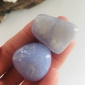 May include: Two smooth, polished blue chalcedony stones. The stones are a soft, pale blue color with subtle white marbling. The stones are held in a hand, showcasing their rounded shapes and natural beauty. These stones are ideal for crystal healing or meditation.