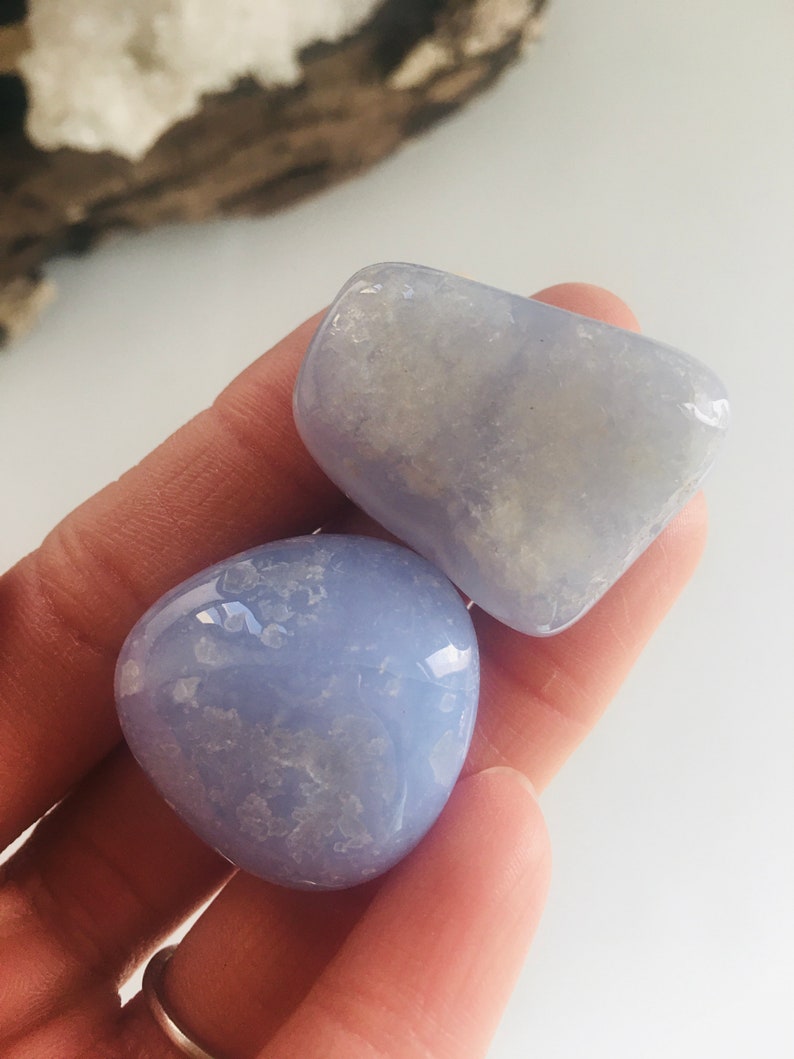 May include: Two smooth, polished blue chalcedony stones. The stones are a soft, pale blue color with subtle white marbling. The stones are held in a hand, showcasing their rounded shapes and natural beauty. These stones are ideal for crystal healing or meditation.