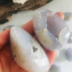 May include: Two smooth, polished blue chalcedony stones with a translucent appearance. The stones have a light blue hue with some areas of white and tan. The stones are held in a hand, with other similar stones visible in the background.