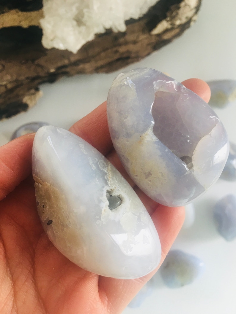 May include: Two smooth, polished blue chalcedony stones with a translucent appearance. The stones have a light blue hue with some areas of white and tan. The stones are held in a hand, with other similar stones visible in the background.