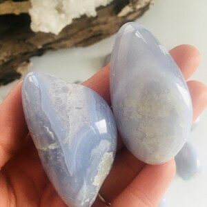 May include: Two smooth, light blue gemstones with white and gray marbling are held in a hand. The stones have a polished, glossy finish. The background includes a piece of driftwood and a white surface.
