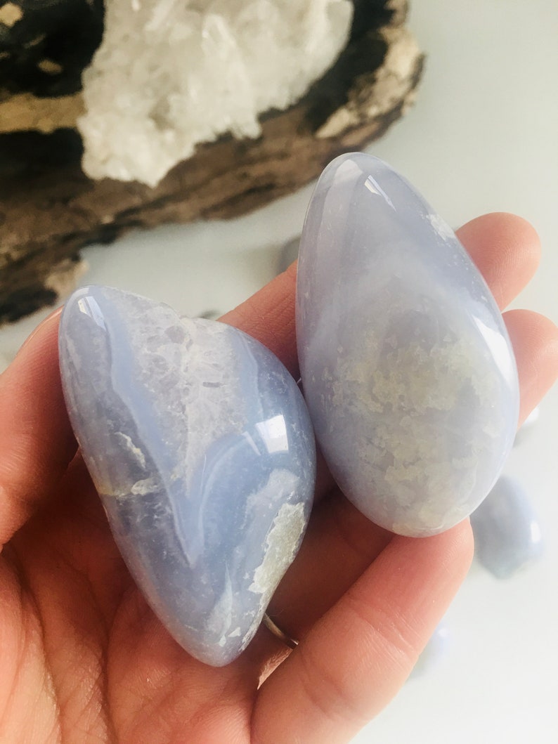 May include: Two smooth, light blue gemstones with white and gray marbling are held in a hand. The stones have a polished, glossy finish. The background includes a piece of driftwood and a white surface.