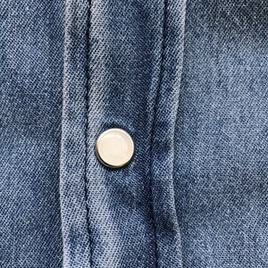 Puede incluir: Primer plano de una camisa vaquera azul con un solo bot&oacute;n redondo nacarado. El bot&oacute;n est&aacute; centrado en la tapeta de la camisa, con costuras visibles y la textura de la tela vaquera.