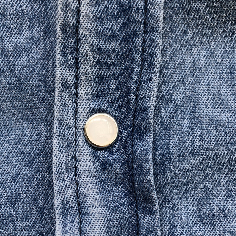 Puede incluir: Primer plano de una camisa vaquera azul con un solo bot&oacute;n redondo nacarado. El bot&oacute;n est&aacute; centrado en la tapeta de la camisa, con costuras visibles y la textura de la tela vaquera.