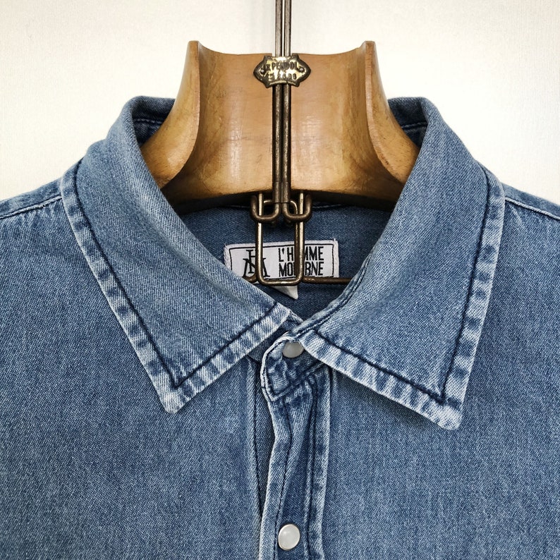 Puede incluir: Una camisa vaquera azul claro con cuello cl&aacute;sico y cierre de botones. La camisa est&aacute; colgada en una percha de madera. Una etiqueta con el texto "L'HOMME MODERNE" es visible cerca del cuello.