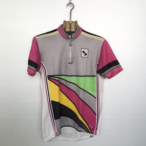 Peut inclure: Un maillot de cyclisme vintage gris, blanc, rose, jaune, noir et vert. Le maillot a une fermeture éclair courte, un petit logo carré et un motif géométrique coloré sur le devant. Les manches sont roses avec des rayures noires et blanches.