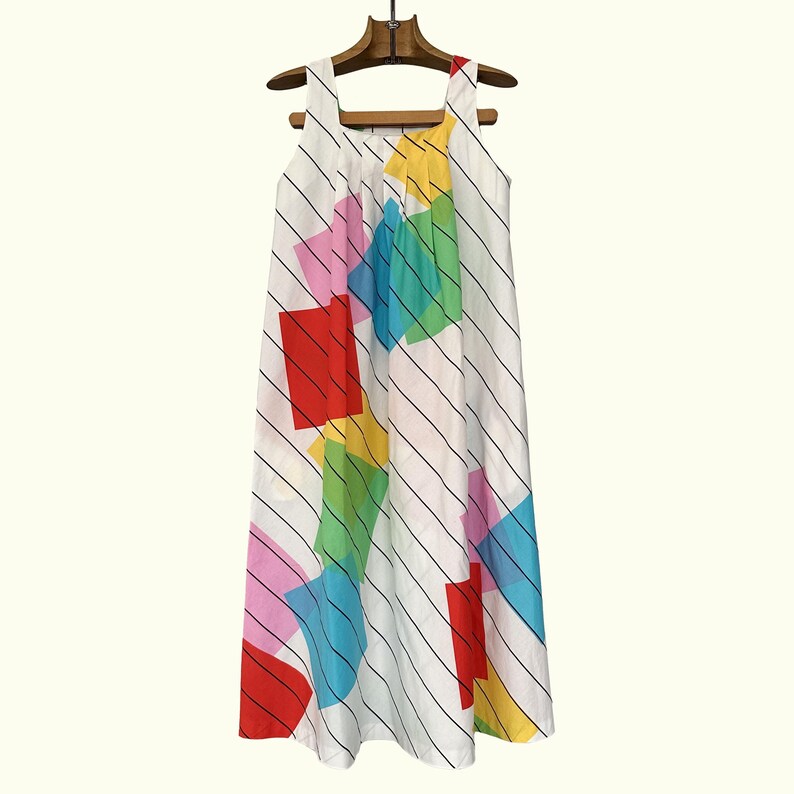 May include: A white sleeveless dress with a vibrant geometric design. The dress has a square neckline and is decorated with large, colourful squares and diagonal black stripes. It is displayed on a wooden hanger.
