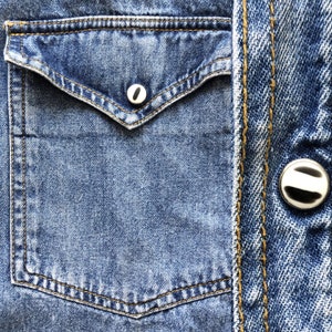Pode incluir: Close-up de um bolso de camisa jeans azul com um bot&atilde;o branco. O bolso tem uma aba pontiaguda e &eacute; costurado com linha amarela. O jeans tem uma apar&ecirc;ncia desbotada e desgastada.