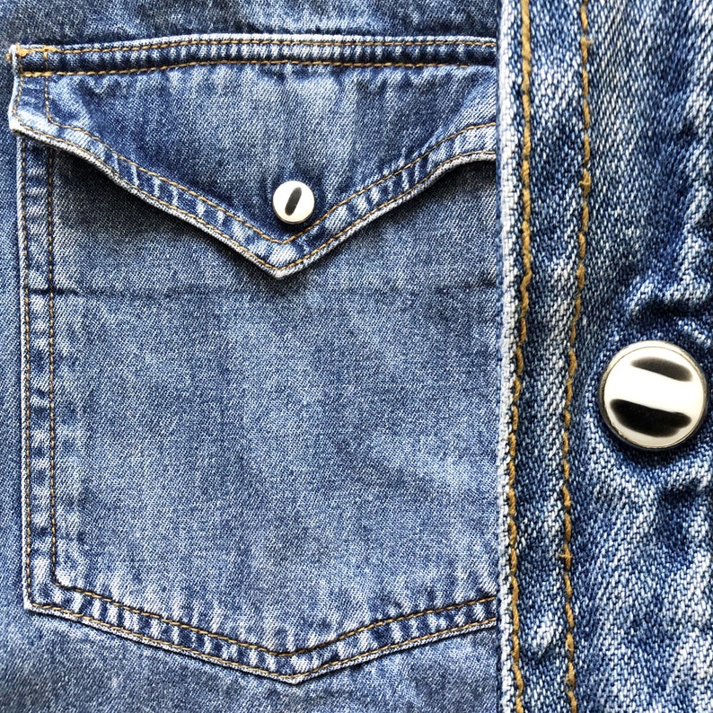 Pode incluir: Close-up de um bolso de camisa jeans azul com um bot&atilde;o branco. O bolso tem uma aba pontiaguda e &eacute; costurado com linha amarela. O jeans tem uma apar&ecirc;ncia desbotada e desgastada.