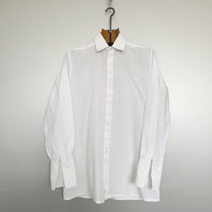 May include: A crisp, white long-sleeve shirt with a classic collar and buttoned cuffs. The shirt is hanging on a wooden hanger against a plain background. The shirt is made of a smooth, likely cotton fabric.