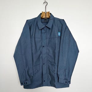 Puede incluir: Chaqueta azul con cierre de botones, dos bolsillos delanteros y un pequeño parche en la manga superior derecha. La chaqueta está colgada de una percha de madera.