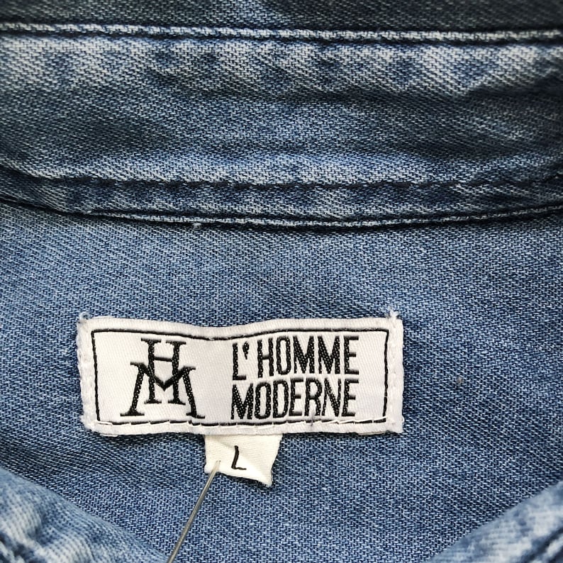 Puede incluir: Primer plano del cuello de una camisa vaquera azul claro. La camisa presenta una etiqueta blanca con el texto "L'HOMME MODERNE" y las iniciales "HM". Tambi&eacute;n es visible una etiqueta de talla "L".