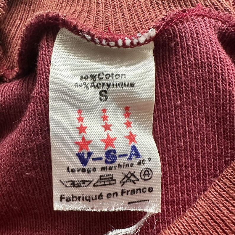 Może przedstawiać: Zbliżenie bordowego materiału z białą metką. Etykieta zawiera tekst, w tym "50% Coton, 40% Acrylique", rozmiar "S" i "Fabriqu&eacute; en France". Etykieta ma r&oacute;wnież czerwone gwiazdki i instrukcje prania.