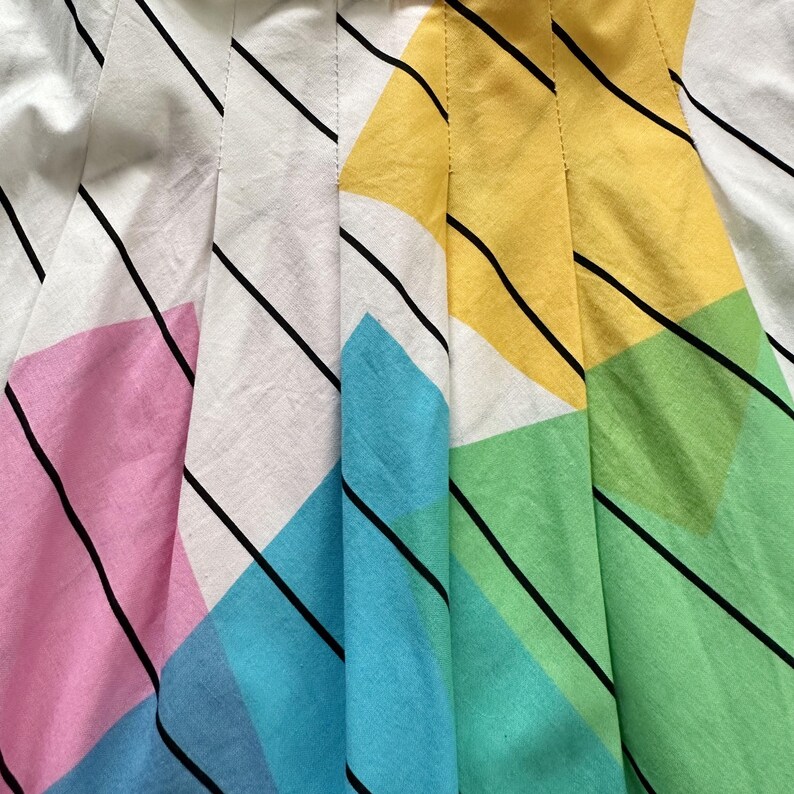May include: Close-up of a pleated fabric with a colourful geometric pattern. The design features white, pink, yellow, blue, and green shapes, accented by black diagonal stripes. The fabric is folded, creating vertical lines.