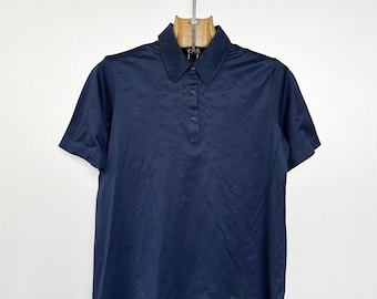 May include: A navy blue polo shirt with short sleeves and a collared neckline. The shirt has a subtle, embroidered floral design on the upper chest. The shirt is hanging on a wooden hanger with a metal hook.