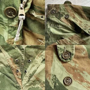 May include: Close-up of a camouflage fabric with multiple buttons and a zipper. The fabric is a mix of green, brown, and tan.