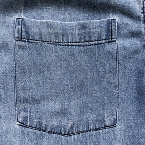 Puede incluir: Primer plano de un bolsillo de camisa vaquera azul claro. El bolsillo es rectangular con una parte superior cuadrada y una parte inferior redondeada. El bolsillo est&aacute; cosido con hilo negro y la mezclilla tiene un aspecto texturizado.
