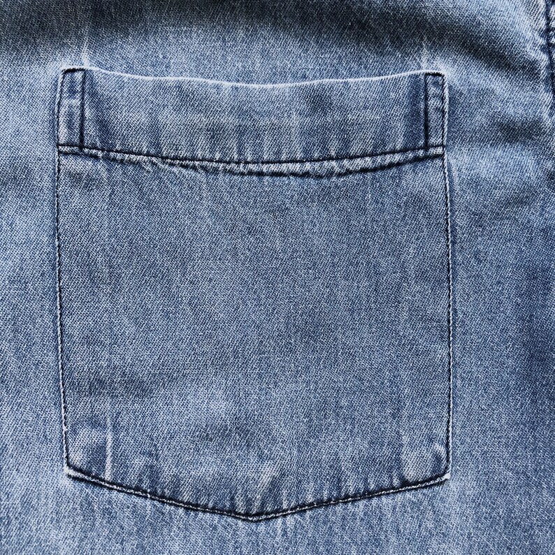 Puede incluir: Primer plano de un bolsillo de camisa vaquera azul claro. El bolsillo es rectangular con una parte superior cuadrada y una parte inferior redondeada. El bolsillo est&aacute; cosido con hilo negro y la mezclilla tiene un aspecto texturizado.