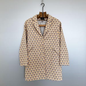 Może przedstawiać: Żakiet w kolorze żółtym i brązowym z geometrycznym wzorem i pojedynczym guzikiem zapięcia. Żakiet ma wycięty kołnierz i dwie kieszenie.