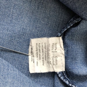Puede incluir: Primer plano de tela vaquera azul claro con una etiqueta blanca. La etiqueta indica "100% Cotton" y "Made in India". Instrucciones de lavado en ingl&eacute;s y franc&eacute;s.