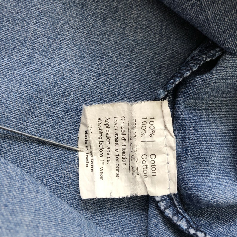 Puede incluir: Primer plano de tela vaquera azul claro con una etiqueta blanca. La etiqueta indica "100% Cotton" y "Made in India". Instrucciones de lavado en ingl&eacute;s y franc&eacute;s.