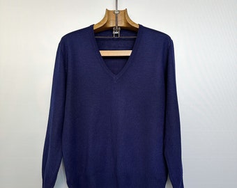 Könnte beinhalten: Ein marineblauer V-Ausschnitt-Pullover mit langen Ärmeln. Der Pullover ist aus einem weichen Strickmaterial gefertigt und hängt an einem Holzkleiderbügel. Der Hintergrund ist eine schlichte weiße Wand.