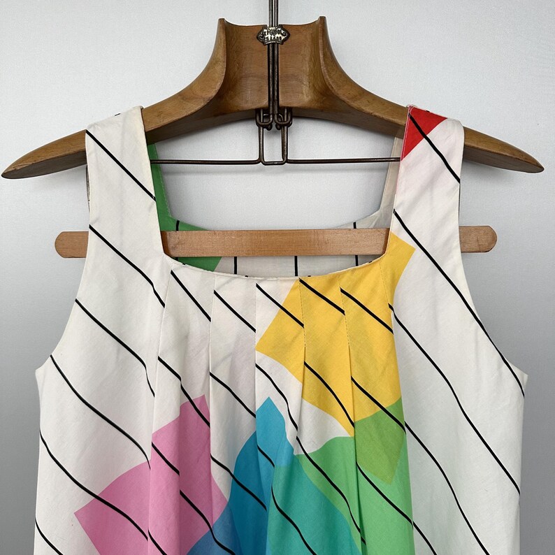 May include: A sleeveless white dress with a colourful geometric design. The dress has a square neckline and is decorated with black diagonal stripes and sections of pink, yellow, green, and blue. It hangs on a wooden hanger.