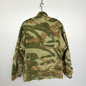 May include: A camouflage jacket with a green, brown, and tan pattern. The jacket has a button closure and two pockets on the front.