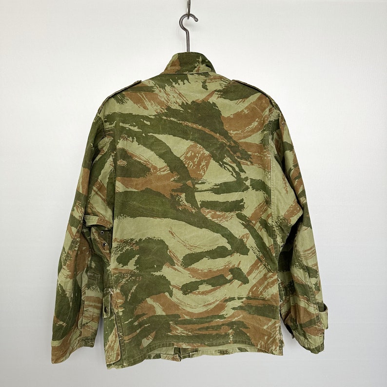 May include: A camouflage jacket with a green, brown, and tan pattern. The jacket has a button closure and two pockets on the front.
