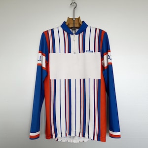May include: A long-sleeved cycling jersey with a white front panel and vertical stripes in red, white, and blue. The sleeves are blue with red accents. The jersey has a half-zip closure and a small logo on the chest.