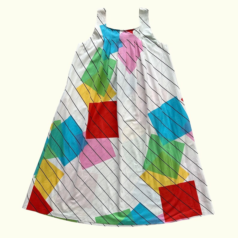 May include: A white sleeveless dress with a colourful geometric design. The dress has diagonal black stripes and bright squares in red, pink, yellow, green, and blue. The dress has a simple, flowing shape.