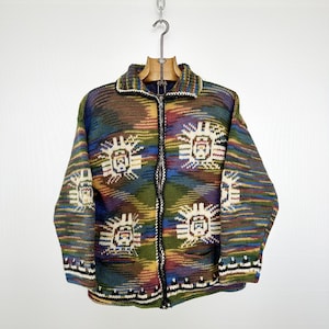 Peut inclure: Un cardigan zippé multicolore avec un motif géométrique. Le pull présente un mélange de couleurs dont le vert, le bleu, le marron et le rouge. Le motif comprend un motif répétitif blanc et beige en forme de soleil. Le cardigan est suspendu à un cintre en bois.