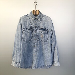 Peut inclure: Une chemise en jean bleu clair avec un devant boutonné et deux grandes poches sur la poitrine. La chemise a un aspect délavé et détérioré.