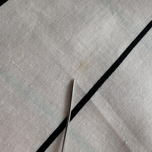 May include: Close-up of a light grey fabric with a black diagonal stripe and a small stain. A silver needle is in the lower portion of the image, pointing towards the fabric.