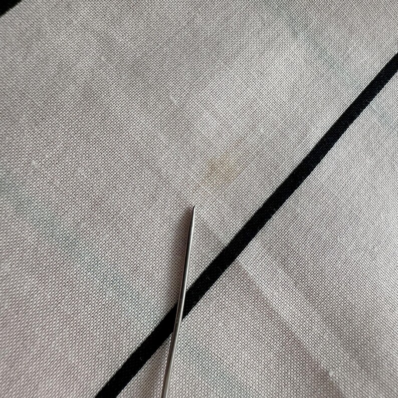 May include: Close-up of a light grey fabric with a black diagonal stripe and a small stain. A silver needle is in the lower portion of the image, pointing towards the fabric.